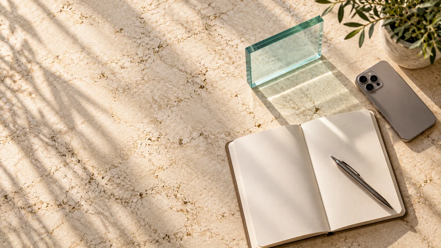 An open notebook with a pen across it, a phone and a single glass sample on a pale stone counter, seen from above in morning light